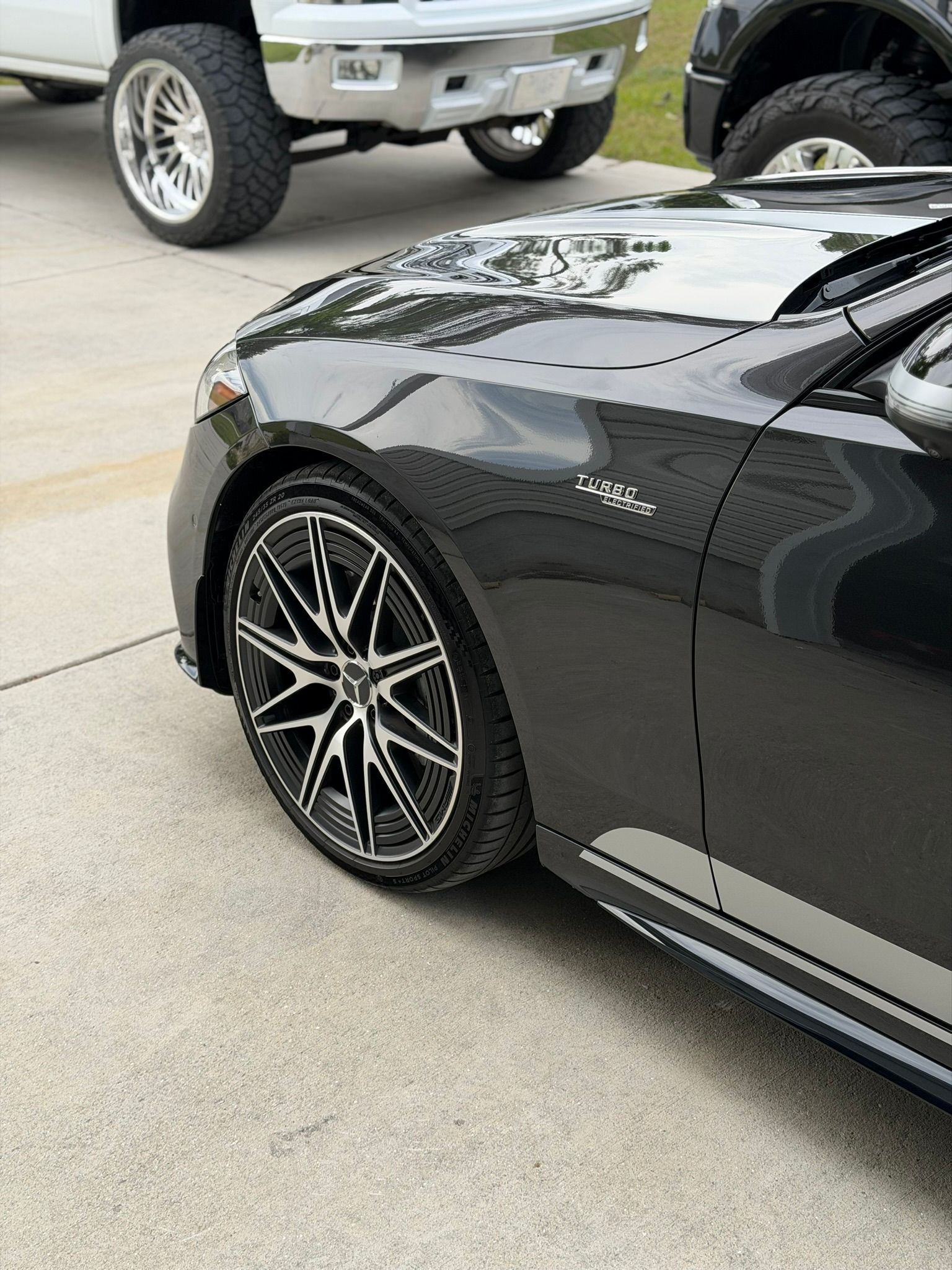 Black sports car front fender and wheel in a driveway, with a silver truck parked behind it