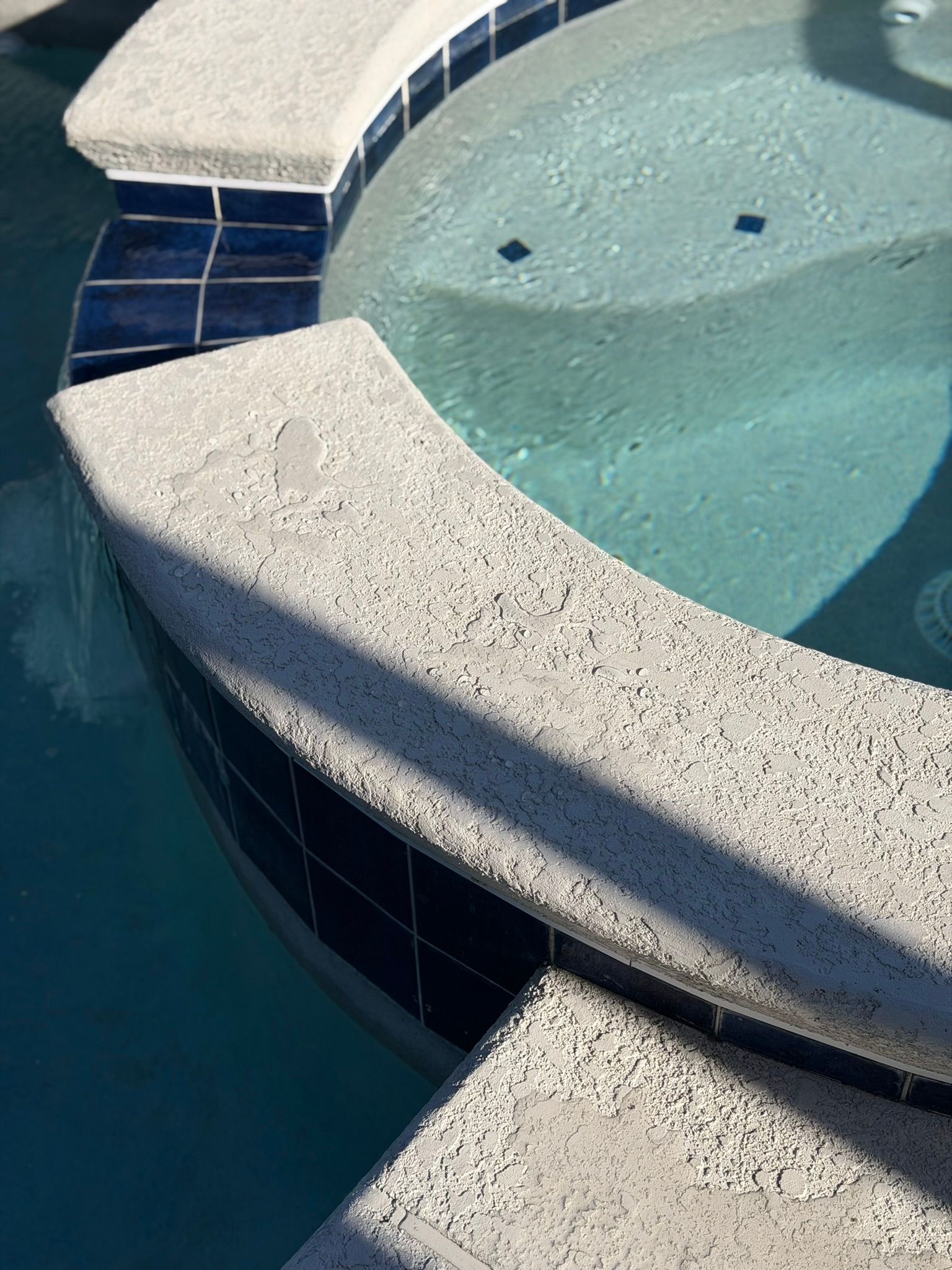Blue-tiled spa pool with curved white stone ledge and light blue water bubbling in sunlight