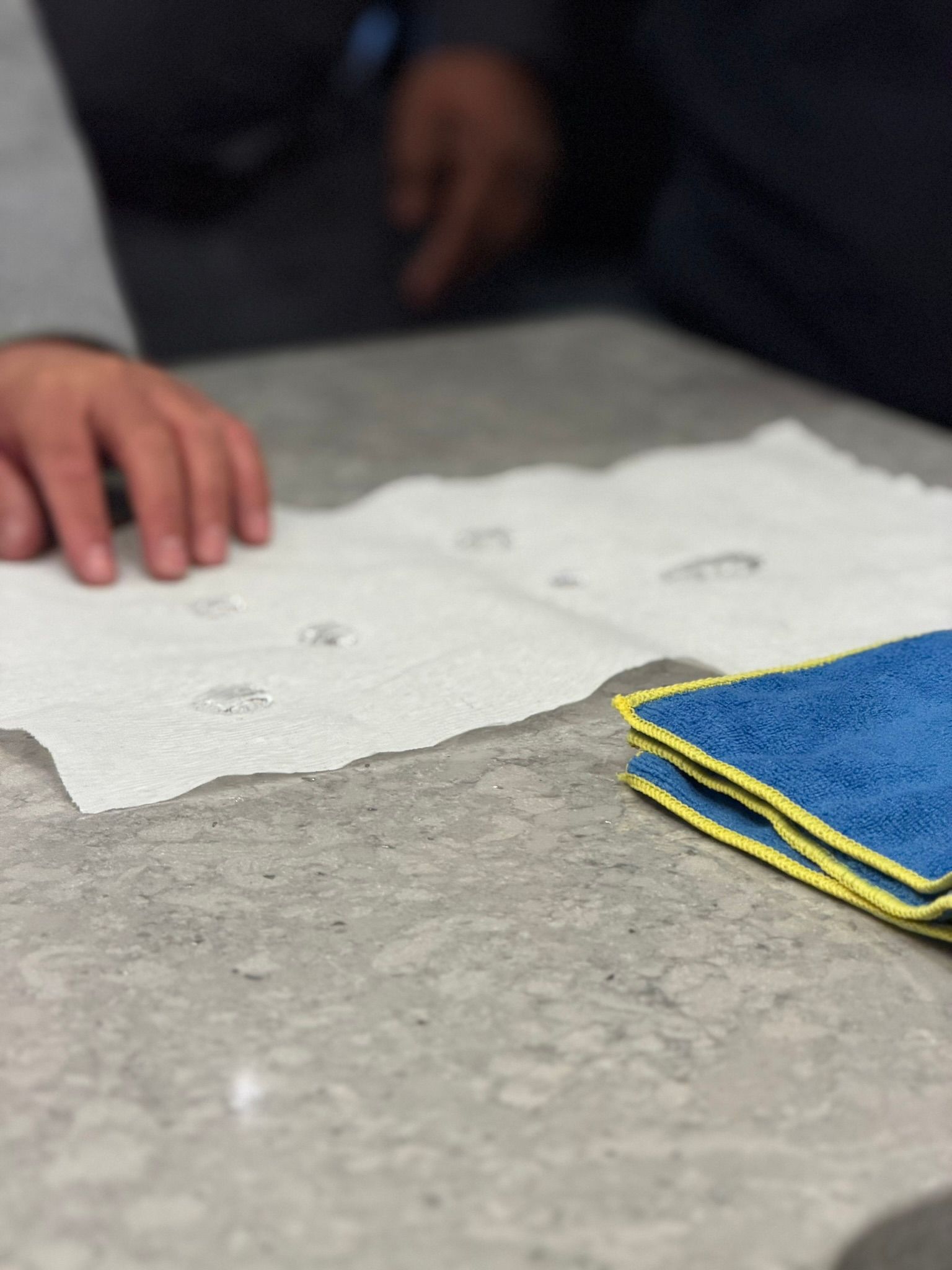 Hands near a gray table with a folded blue and yellow cloth on the right.