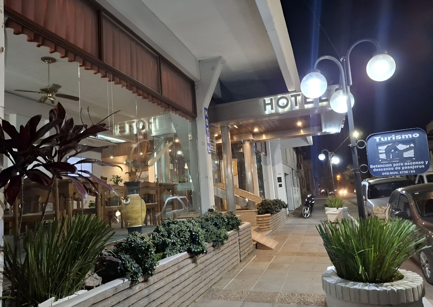 Entrada nocturna del hotel con faroles luminosos, plantas en macetas y un pasillo embaldosado.