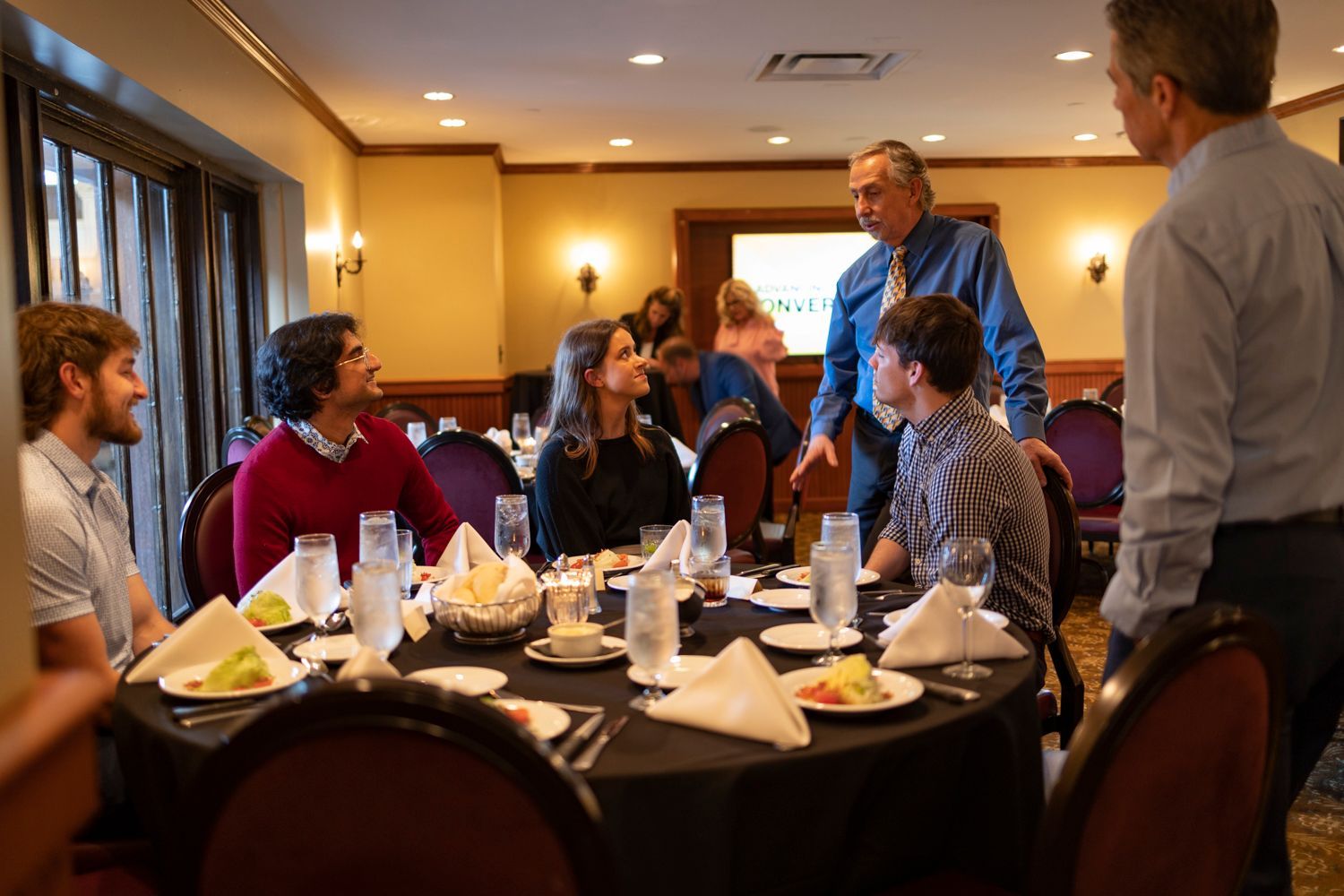 A group of people are sitting at a table in a room talking to each other.