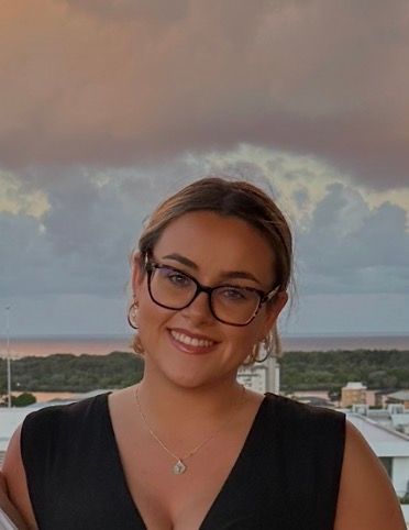Smiling woman in glasses and a black top standing outdoors at sunset with cloudy sky behind her