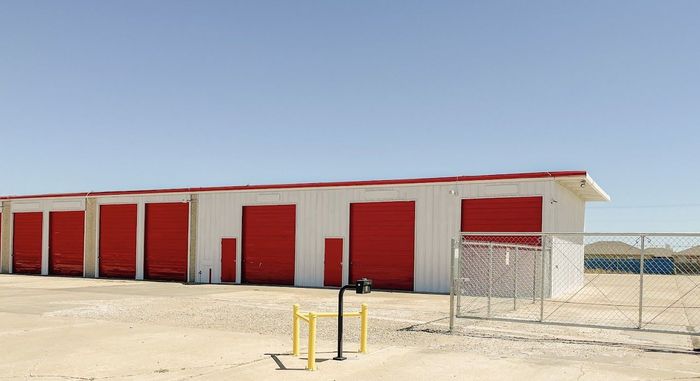 Row of red storage unit doors on a white building in a sandy, desert-like lot under a clear blue sky