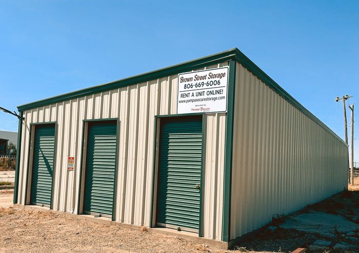 Small beige metal storage building with green doors in a gravel lot under a clear blue sky