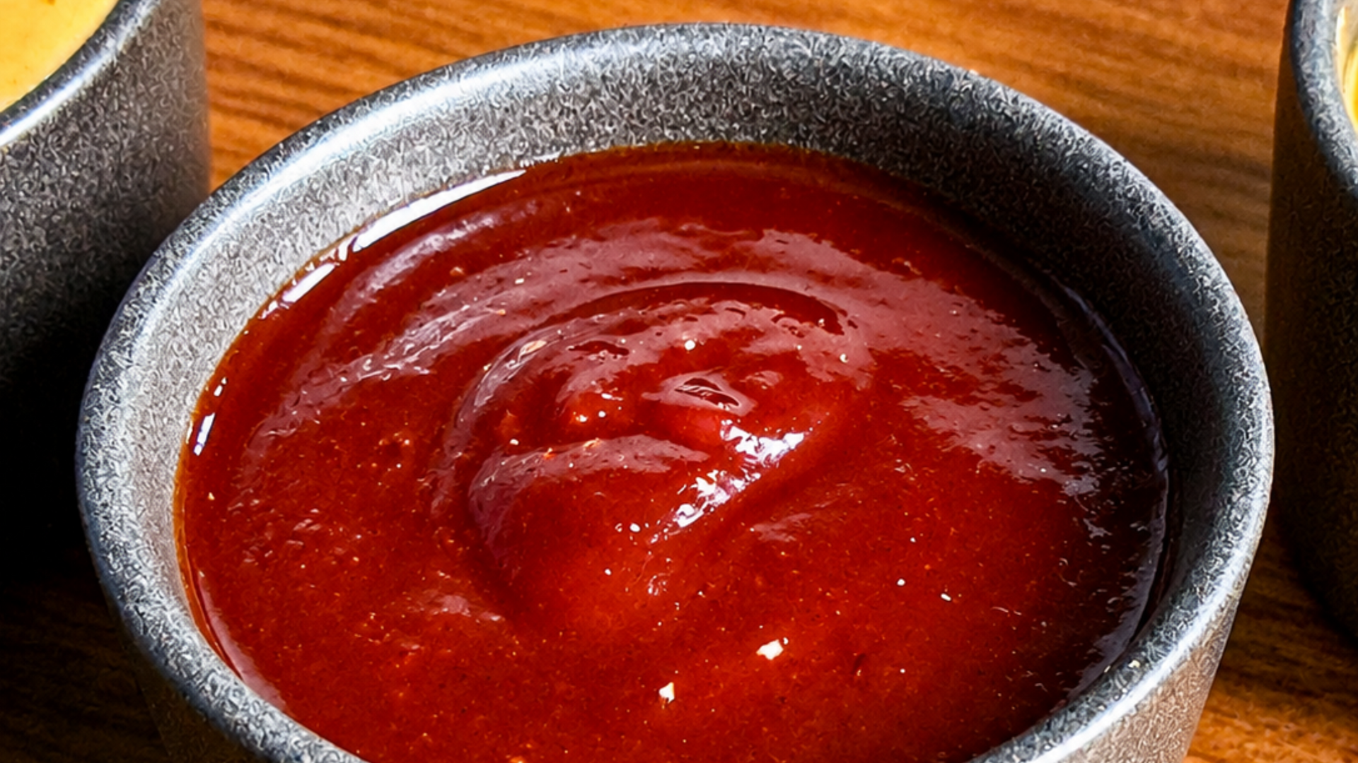 A bowl of glossy red sauce or soup on a wooden table, with another bowl partially visible at right.