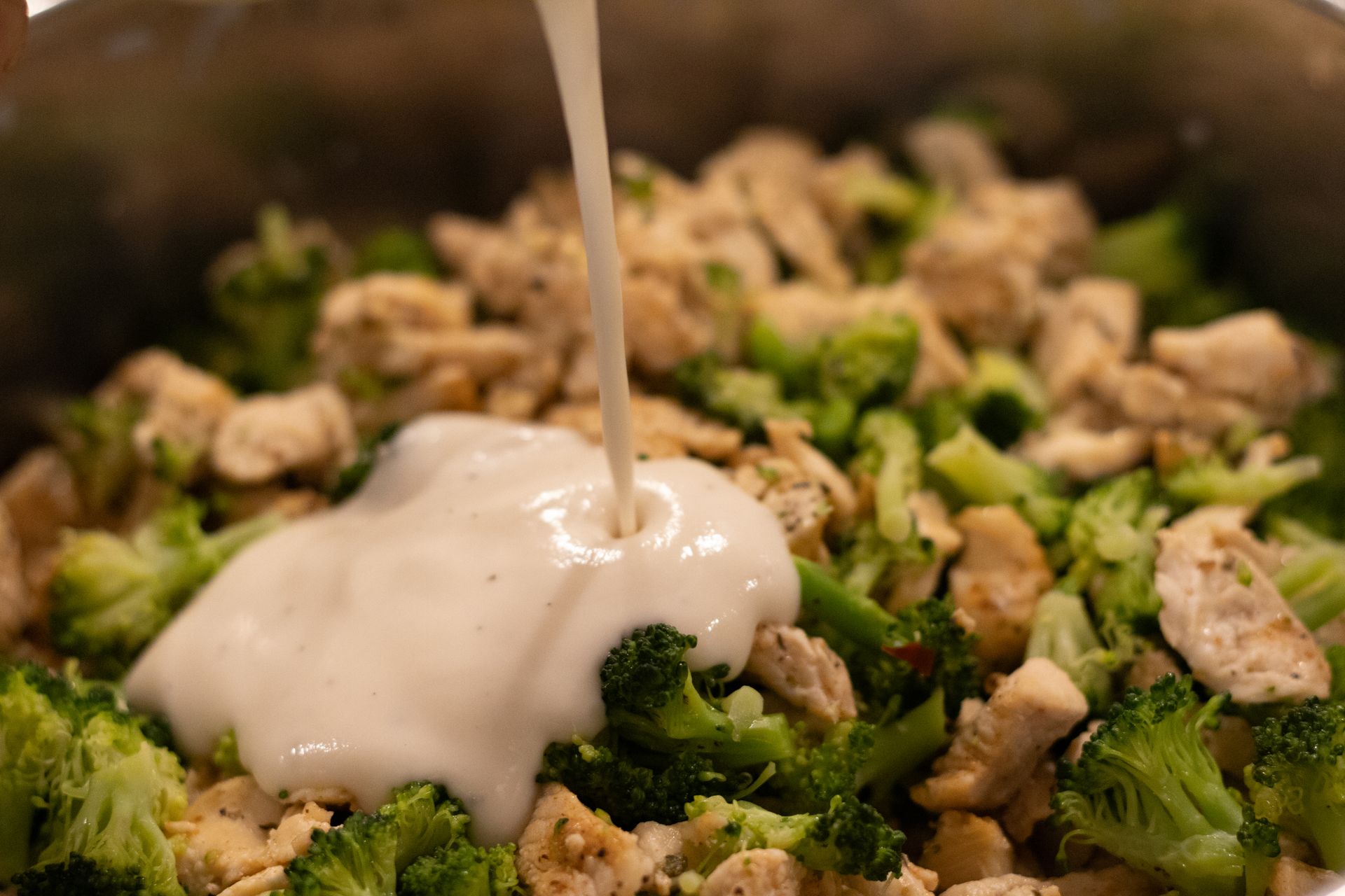 A person is pouring sauce into a pot of broccoli and chicken.