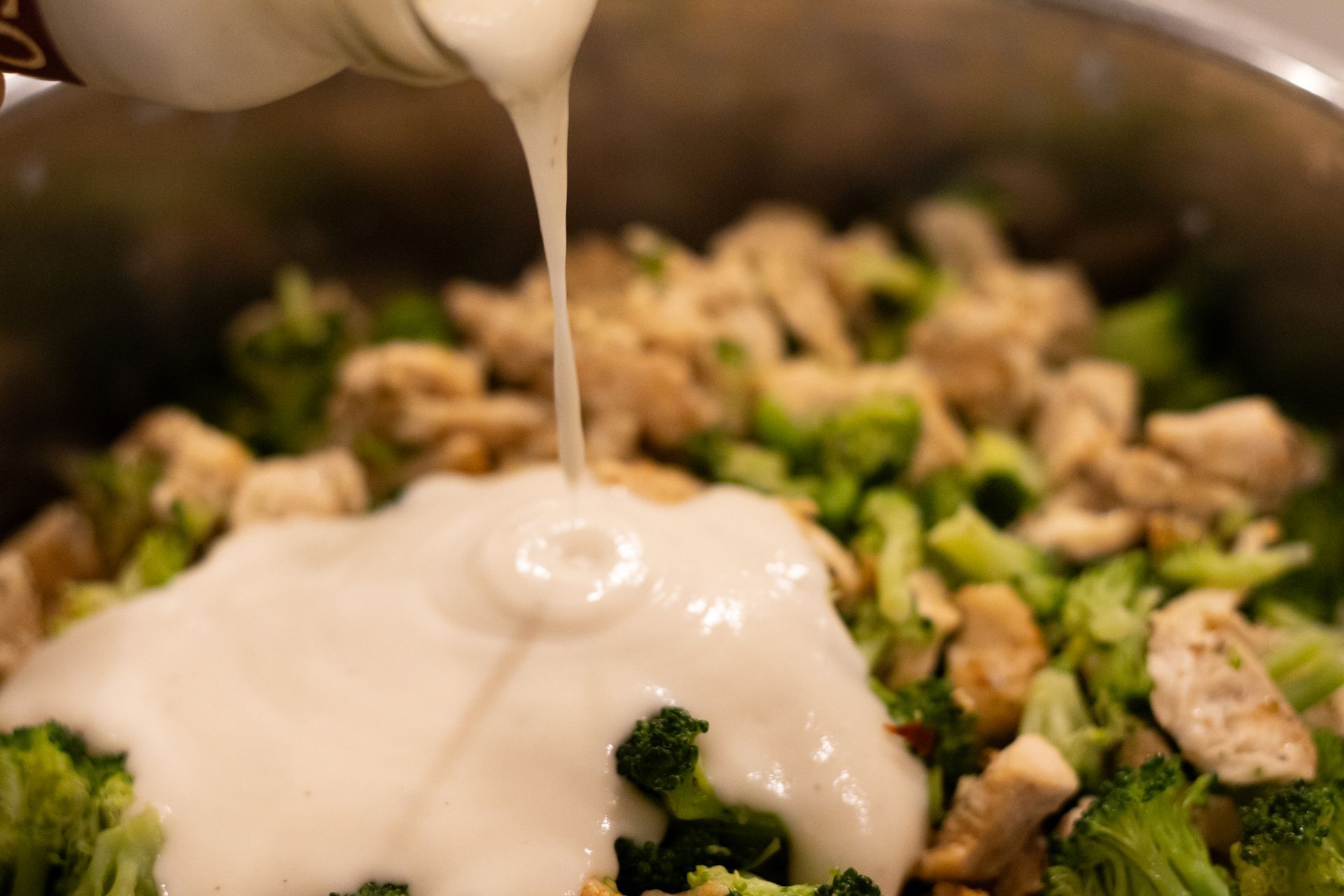 A person is pouring sauce into a pot of broccoli and chicken.