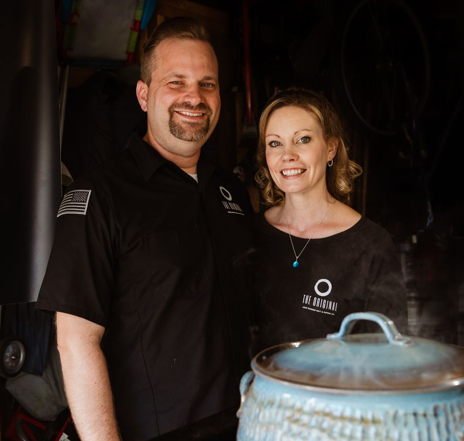 Two people in black shirts smile beside a large metal pot in a dim kitchen or bar area.