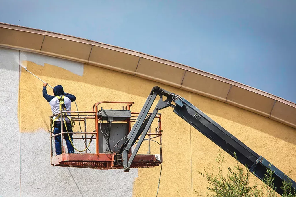 Worker in a lift painting a tan building wall under a blue sky