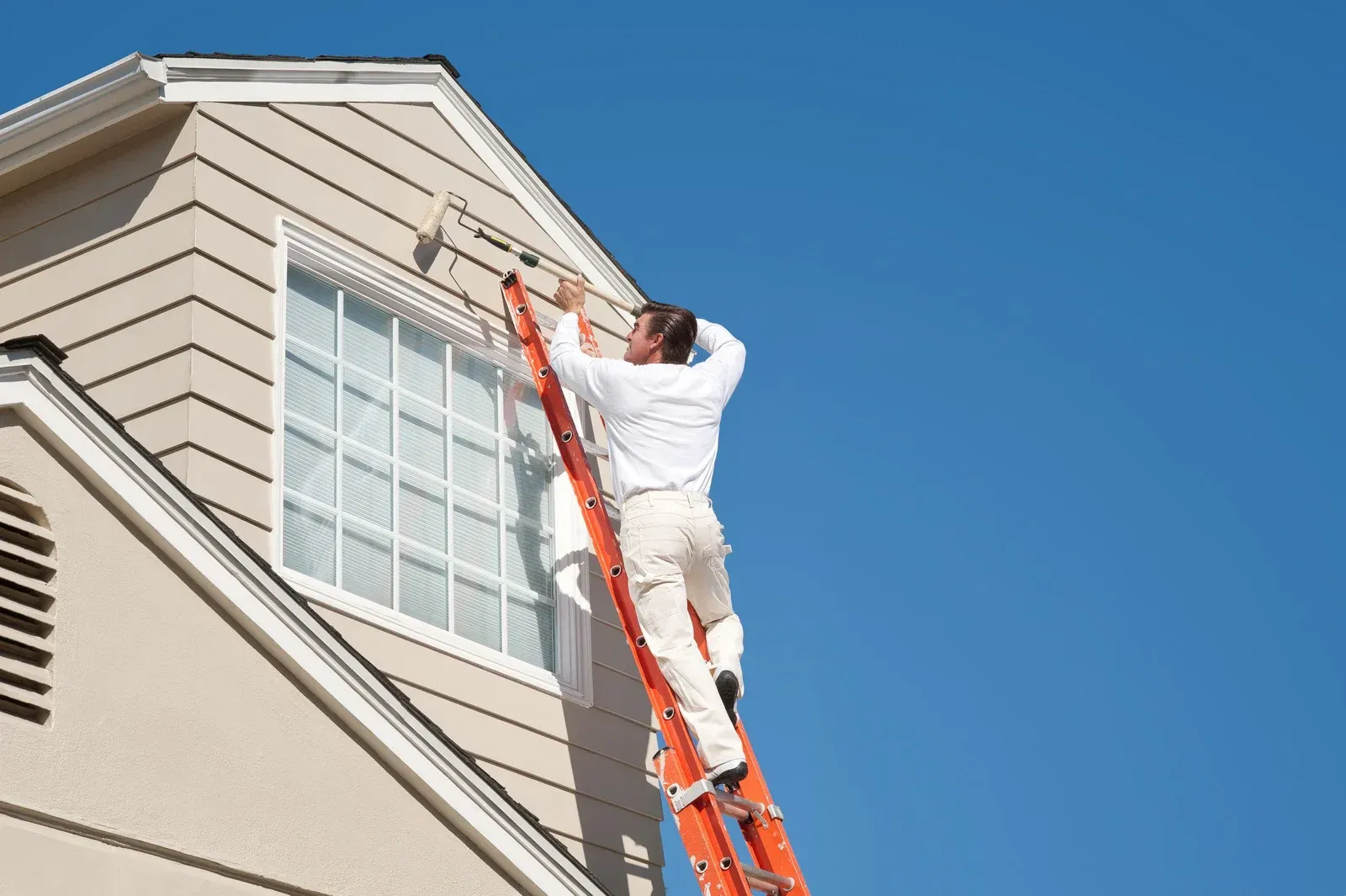 Worker on orange ladder painting the trim of a beige house under a clear blue sky