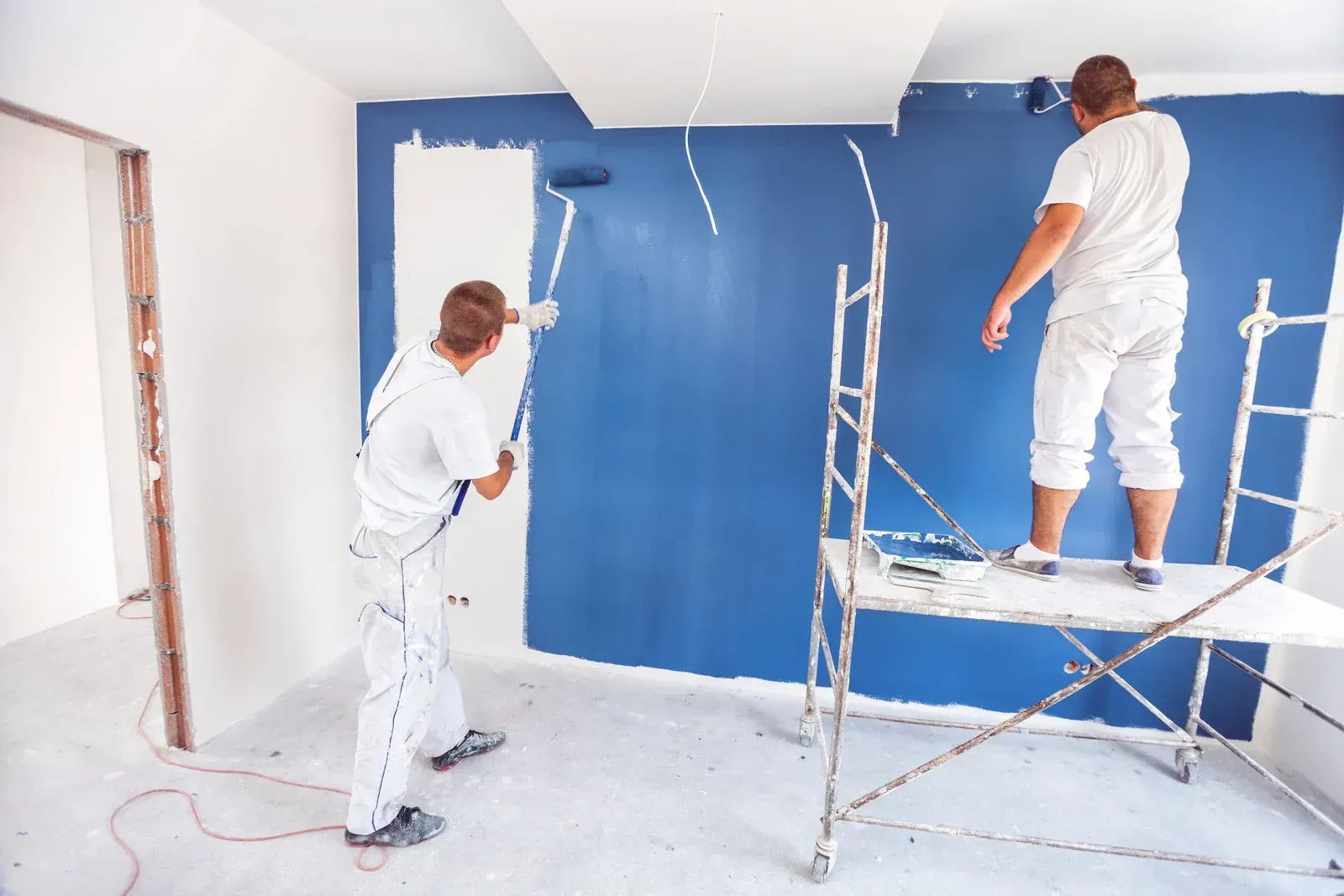 Two workers painting a room blue, one on scaffolding and one using a roller