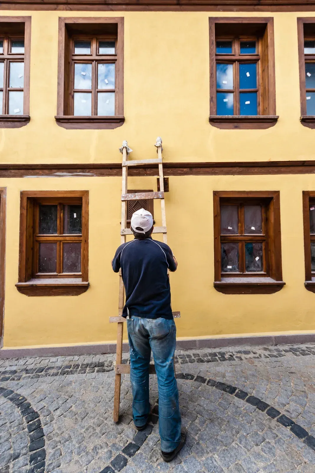 Person standing on a cobblestone street facing a yellow building with wooden windows and a ladder