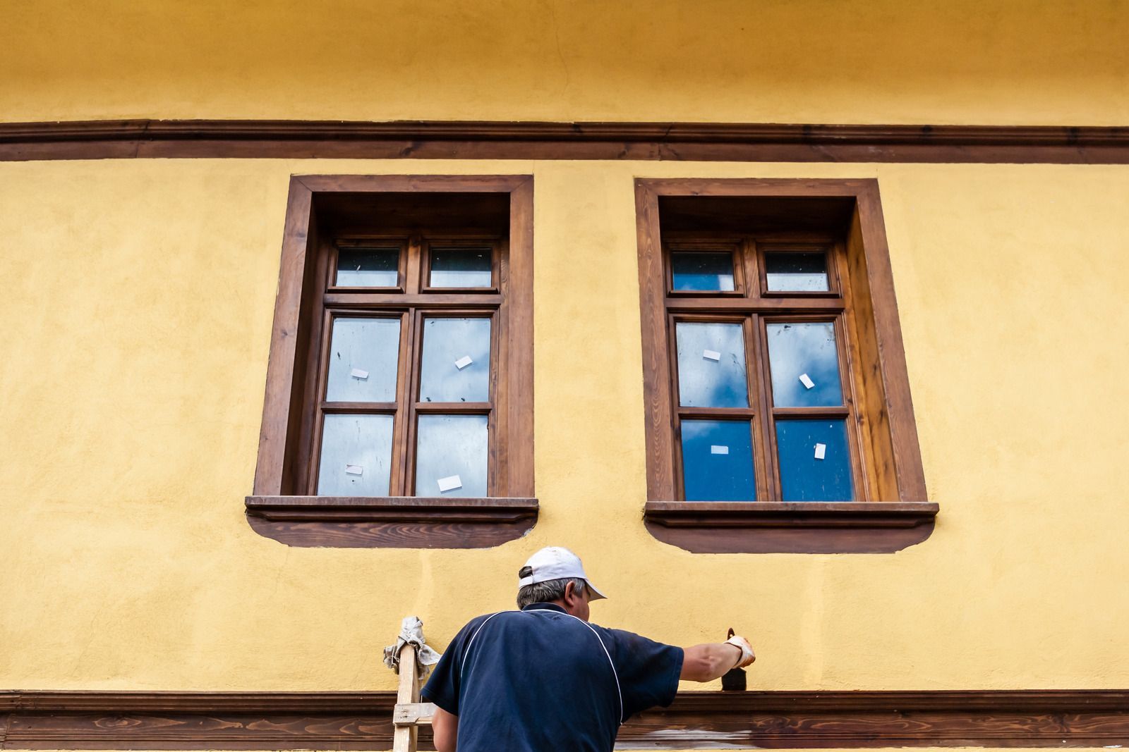 Person in a hat painting a yellow wall beneath two wooden-framed windows