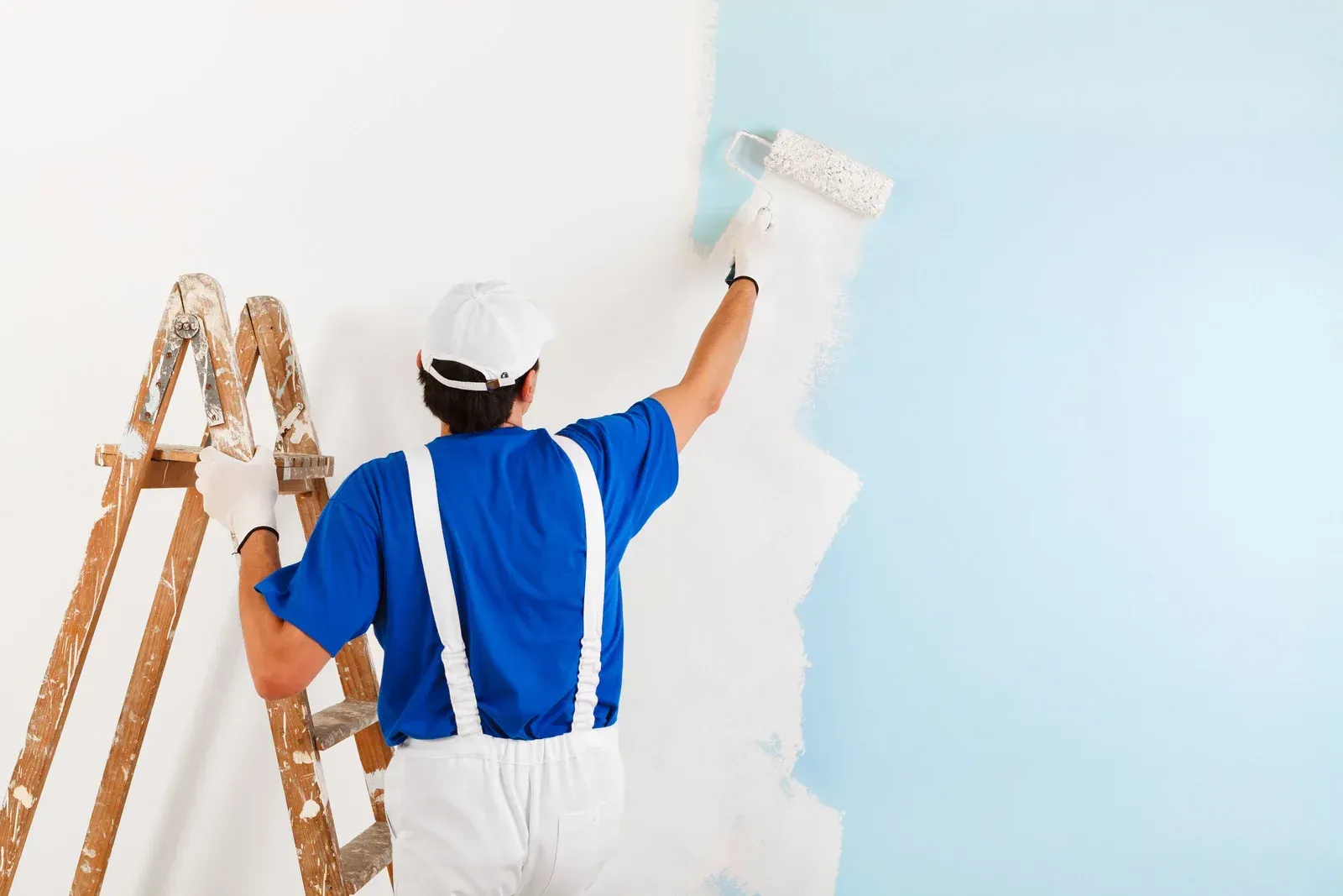 Painter in blue overalls painting a white wall next to a wooden ladder