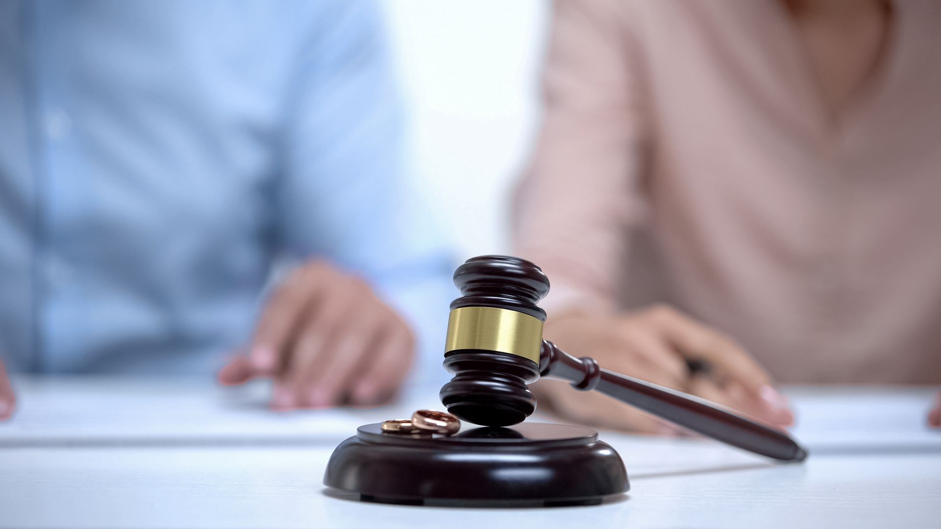 Divorce attorney gavel and wedding rings on desk with couple reviewing separation documents. Divorce attorney gavel and wedding rings on desk with couple reviewing separation documents.