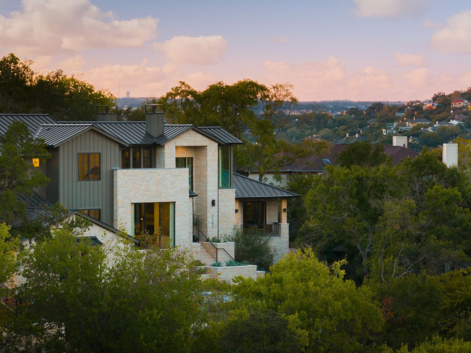 House with light stone facade, green siding, and trees at dusk. Distant cityscape in background.