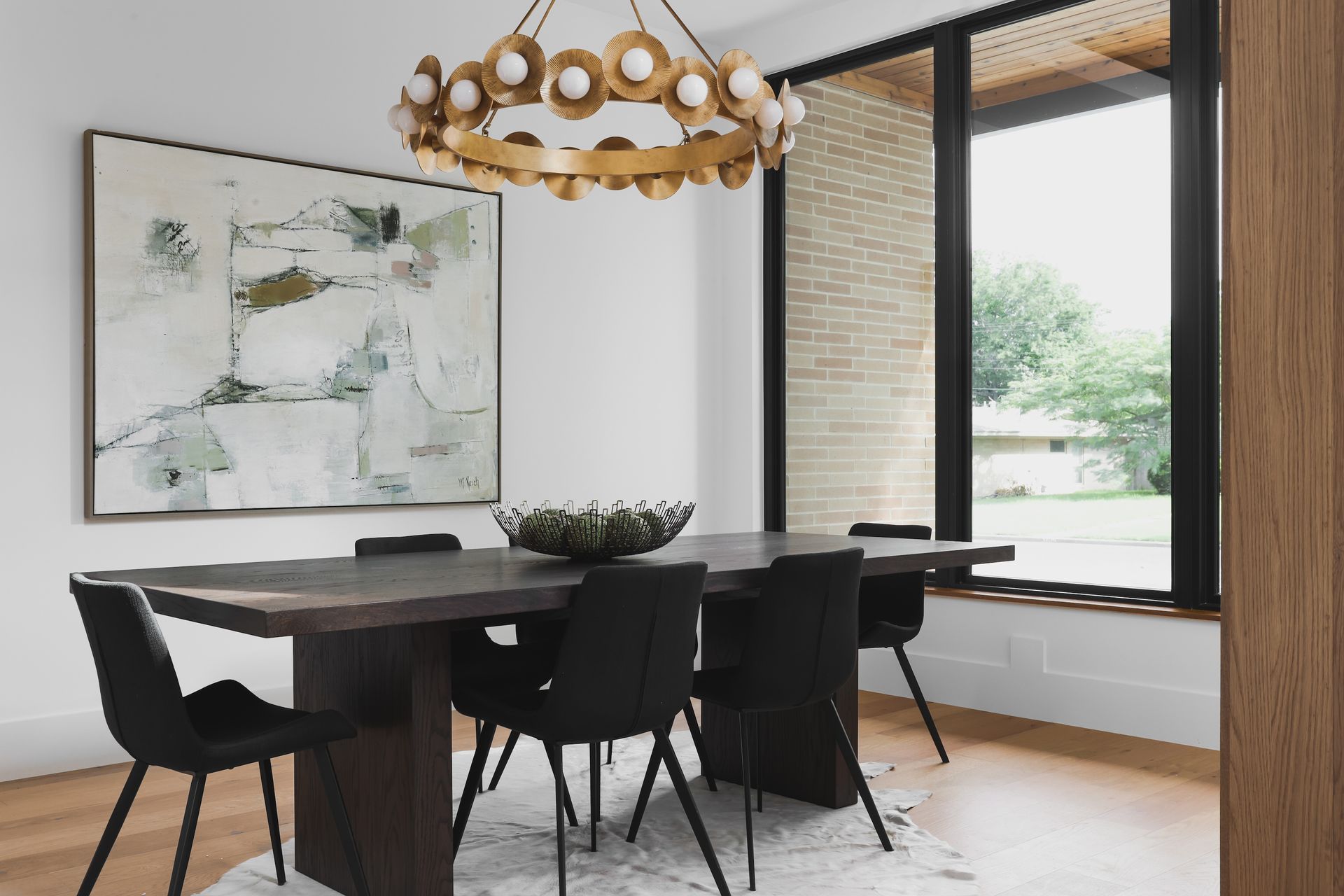 Dining room with dark wood table, black chairs, and gold chandelier. Large abstract art and window.