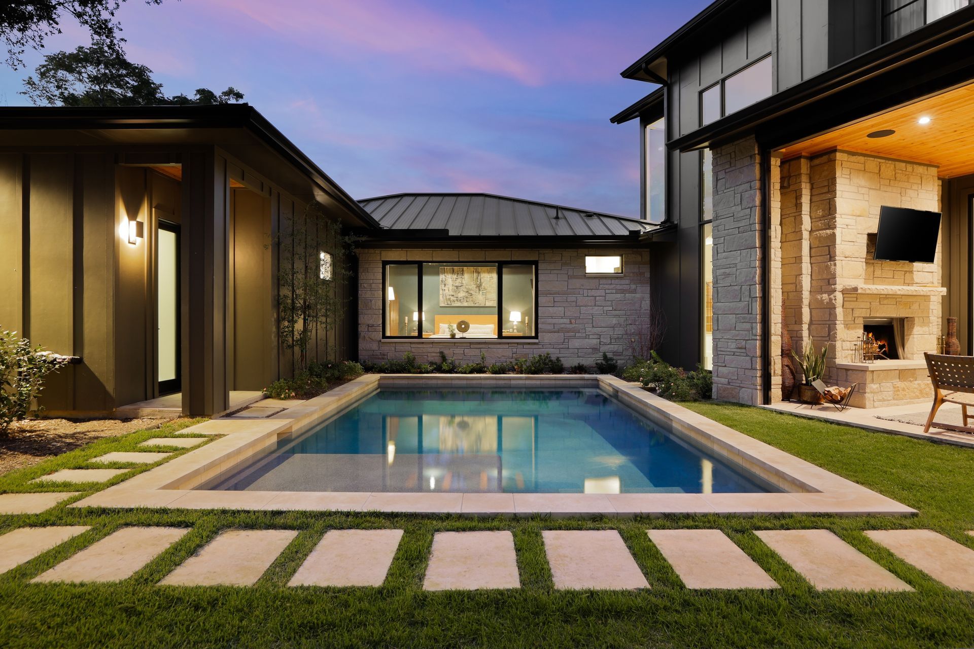 Backyard with a rectangular pool, surrounded by grass and stone pavers, between two buildings, with a fireplace.