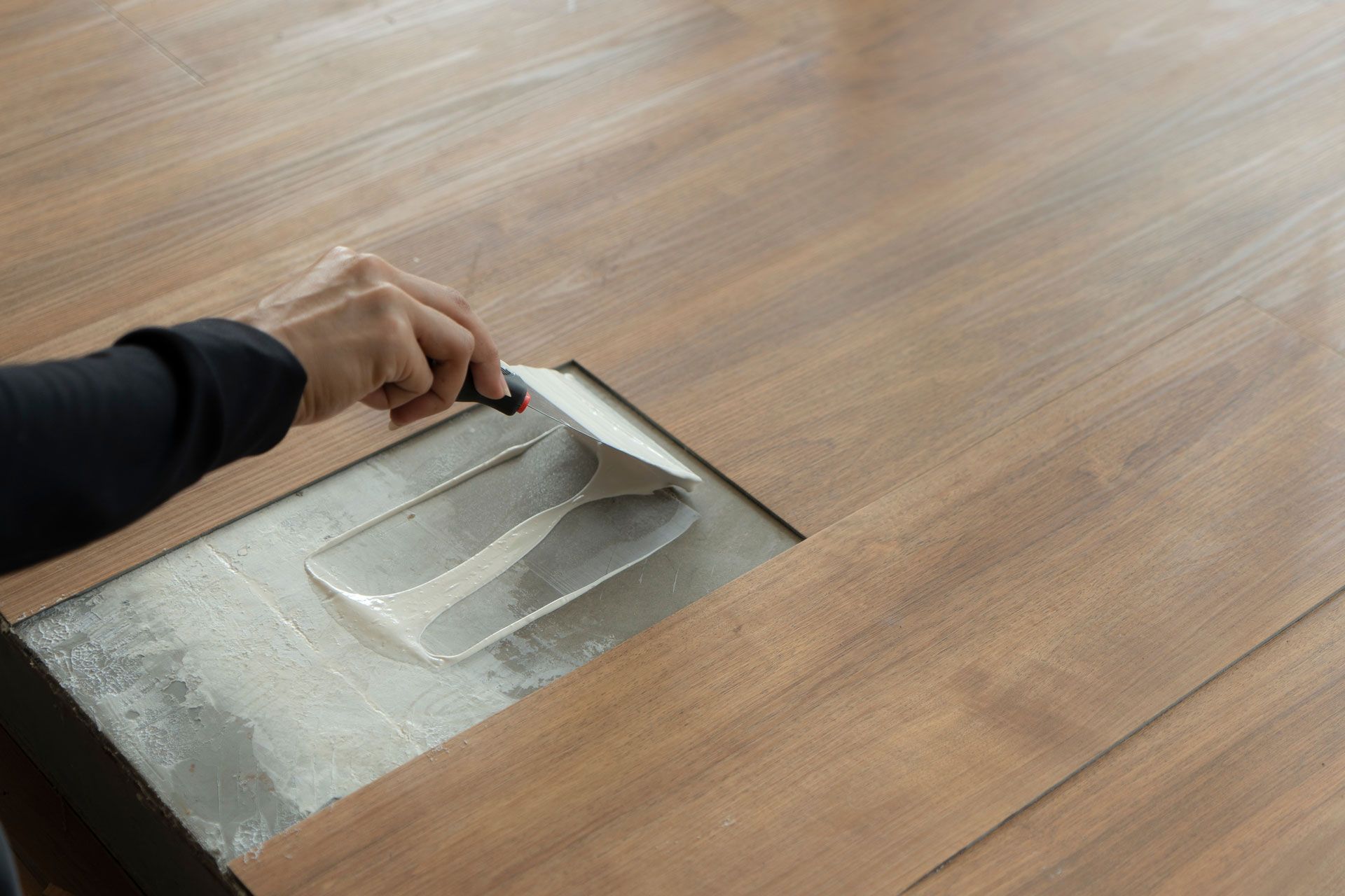 Hand placing a metal trowel on a wood-look floor tile beside an open square cutout
