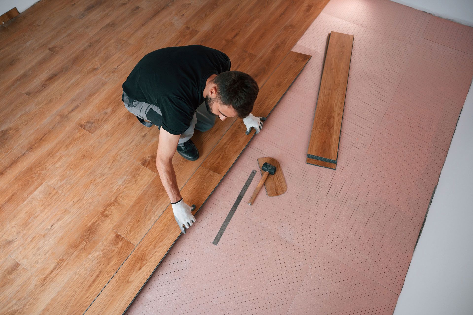 Worker installing wooden flooring beside a pink carpet, using tools on the floor