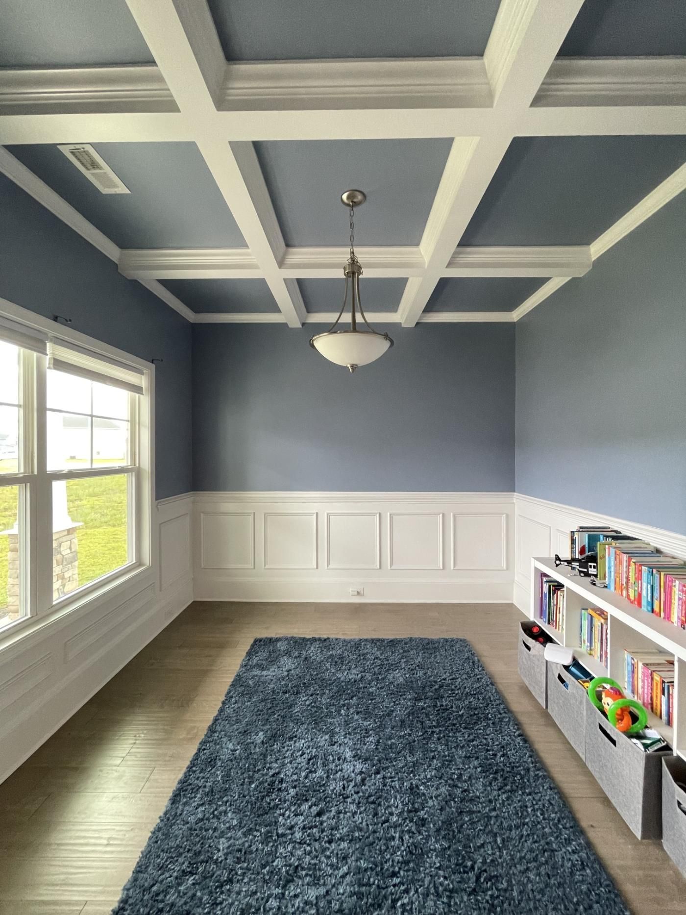 Empty room with blue-gray walls, white wainscoting and coffered ceiling, window, rug, and bookshelf.