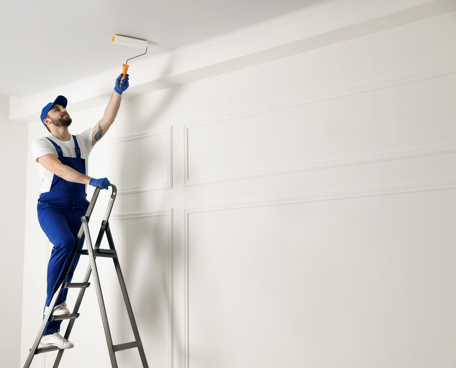 Worker on a ladder painting a white ceiling with a roller in a bright room