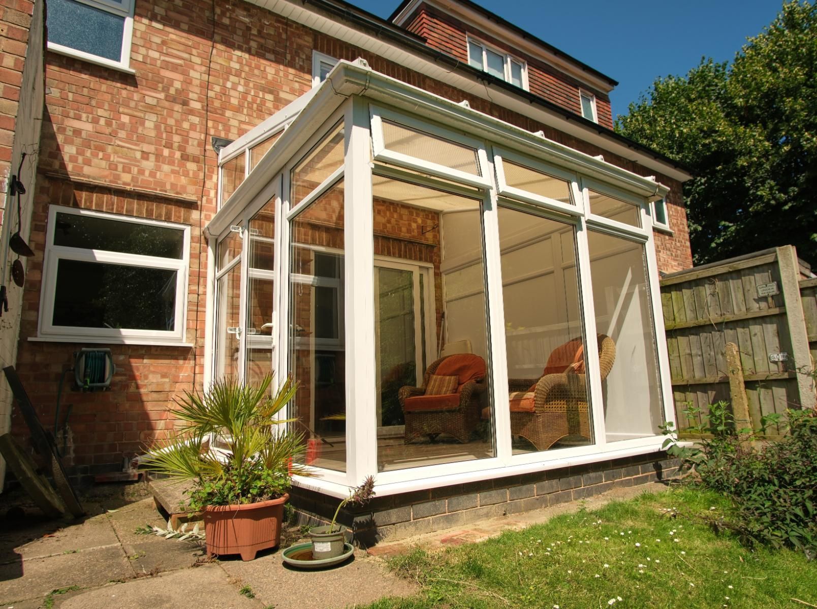 Backyard glass sunroom attached to a brick house, with chairs inside and potted plants outside.