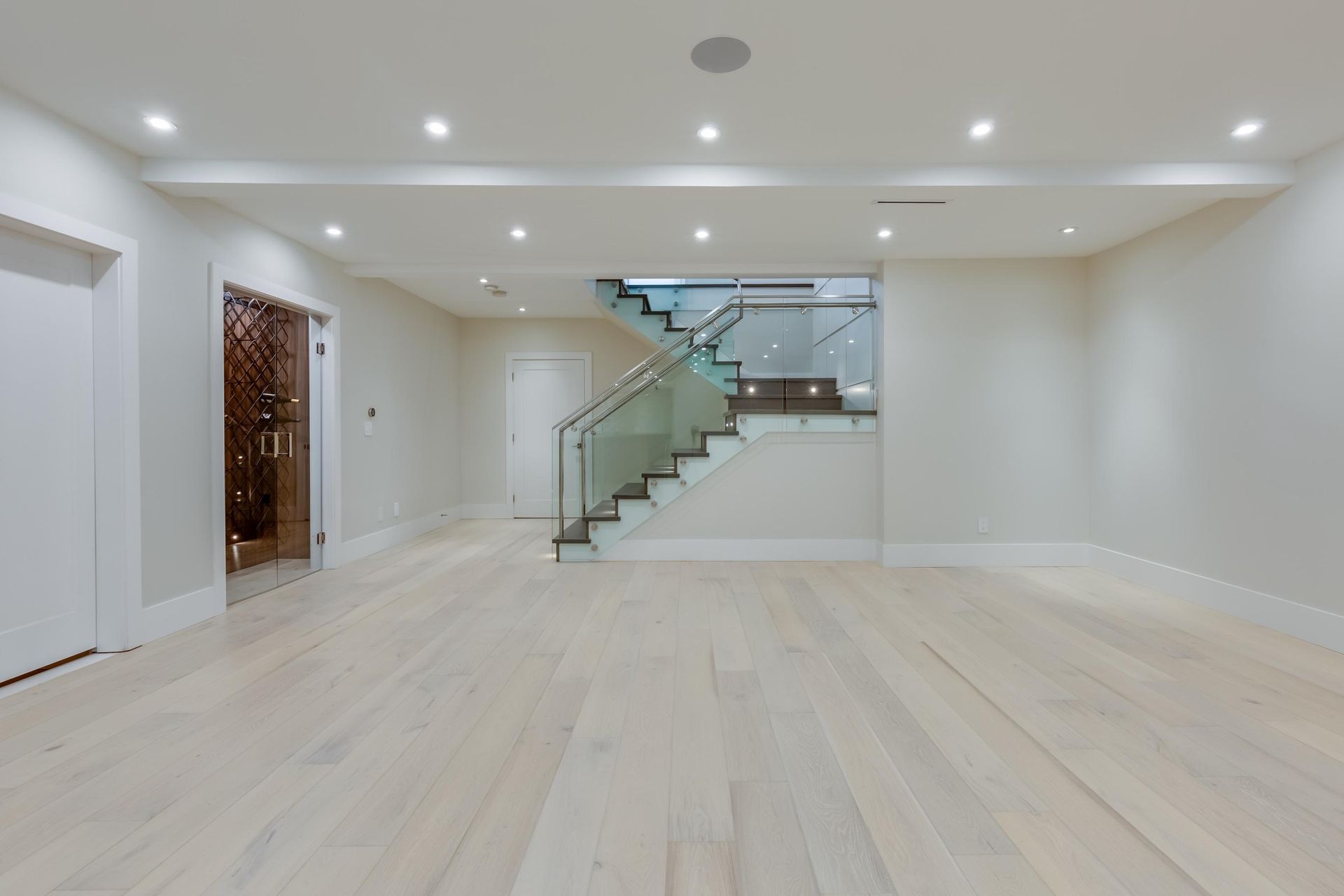 Empty modern living room with light wood floors, white walls, recessed lights, and a glass staircase.