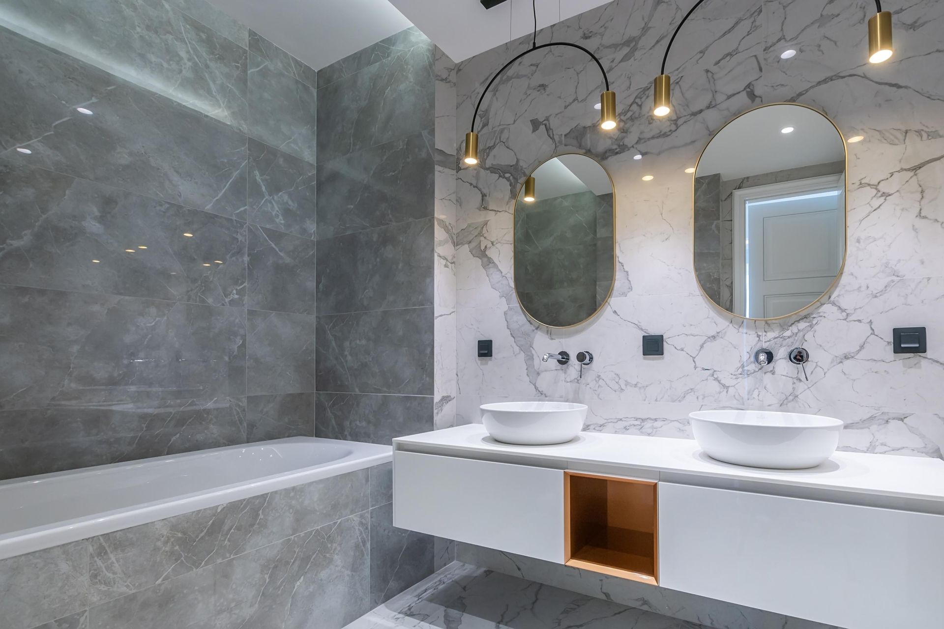 Modern bathroom with gray marble walls, double vanity, oval mirrors, and a bathtub on the left