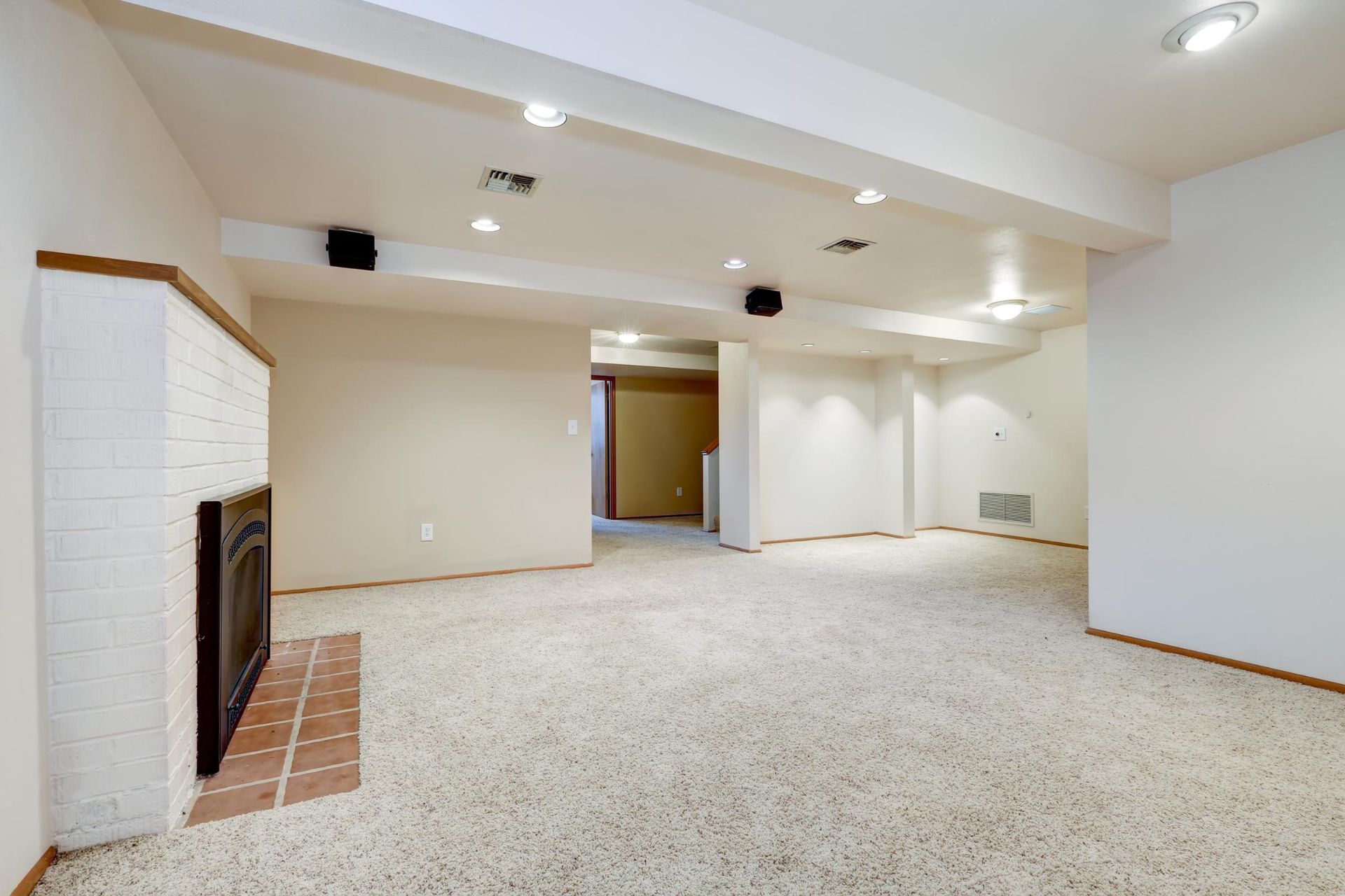 Empty carpeted basement rec room with white walls, recessed lights, and a fireplace on the left.