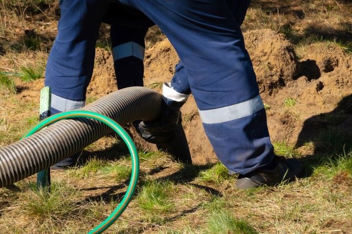 Person in blue overalls inserting a large hose into a hole in the ground.