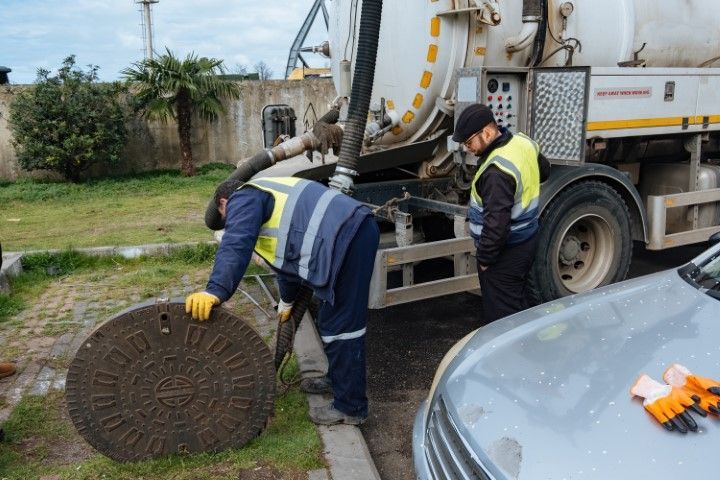 Two workers in vests, one opening a sewer, another near a waste truck, outdoors.