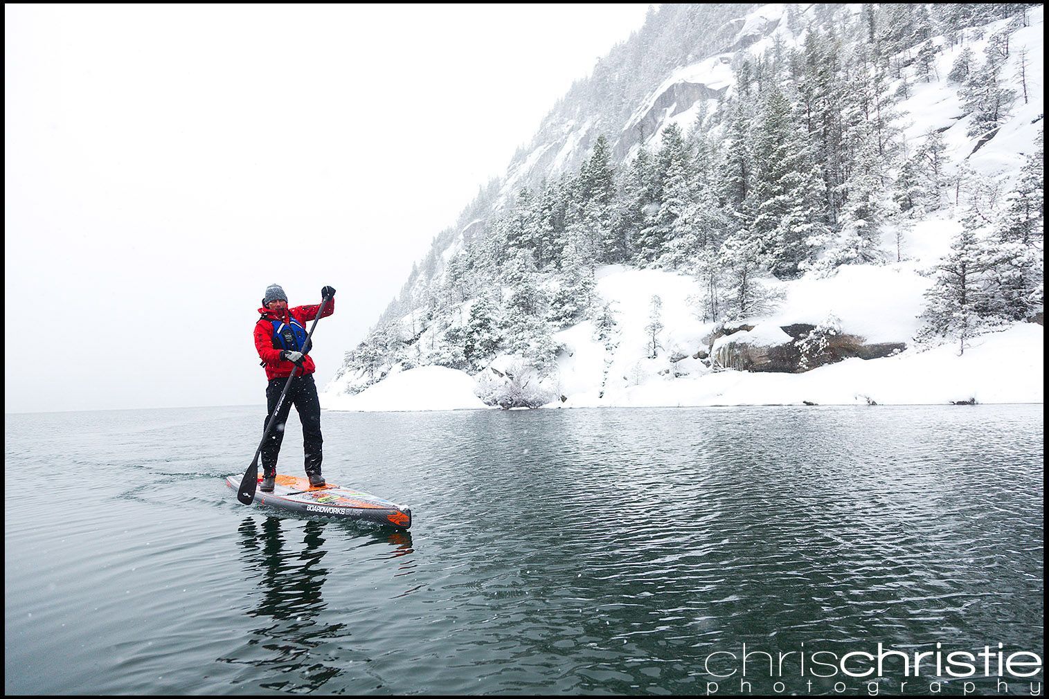 Person in red jacket paddleboarding on a snowy, icy lake beside a steep winter mountain