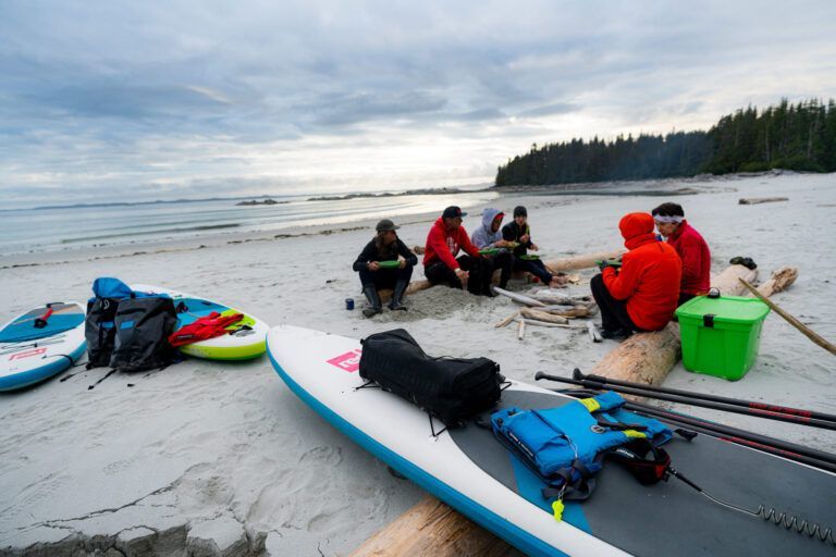 People resting on a sandy beach beside paddleboards and kayak near calm water and a forested shoreline