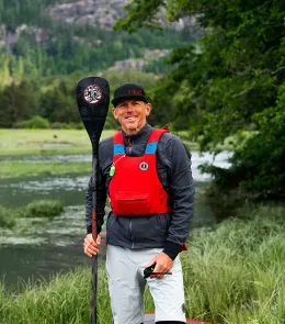 Smiling kayaker in red life vest holds paddle beside a river with mountains and trees in the background