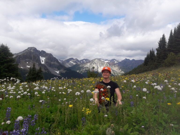 Person in an orange cap sitting in a wildflower meadow with mountains and cloudy sky behind.
