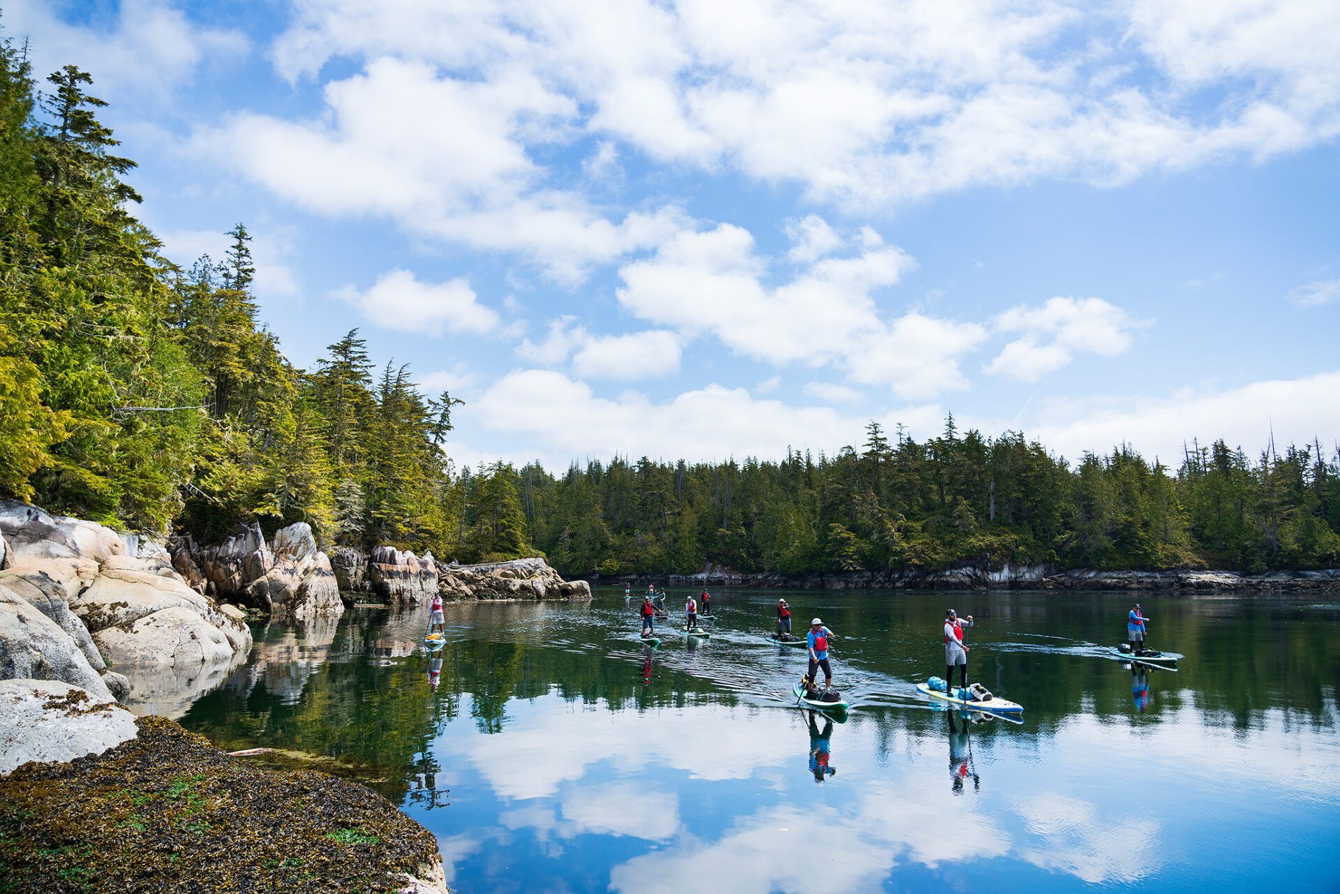 Kayakers paddling on a calm forest lake with rocky shores and blue sky reflections