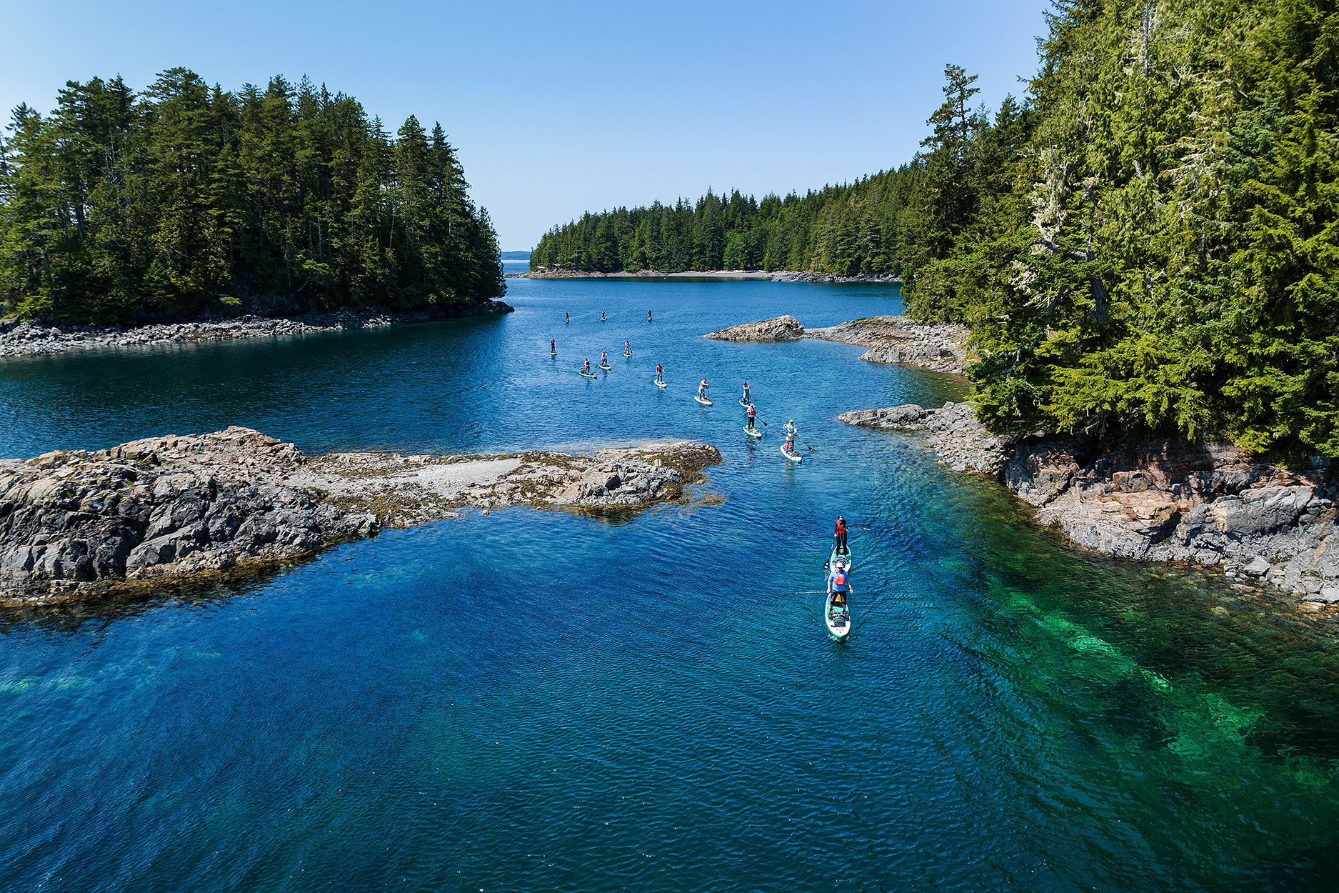 Kayakers in a narrow turquoise channel between rocky, tree-lined islands under a blue sky