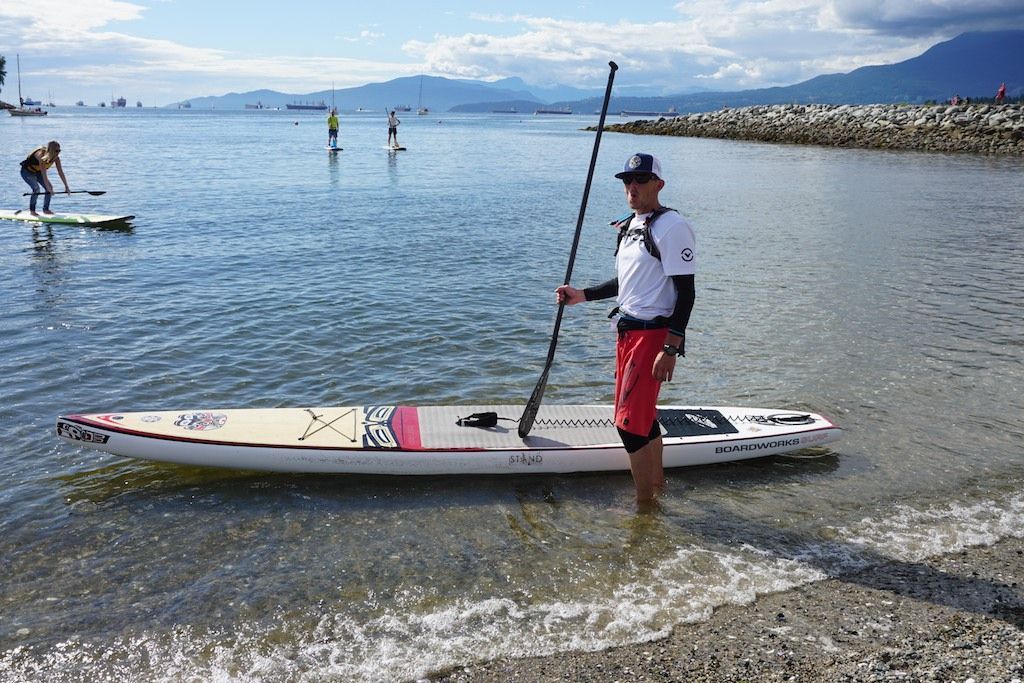 Person stands beside a paddleboard in shallow water on a rocky beach, with people paddling in the distance.