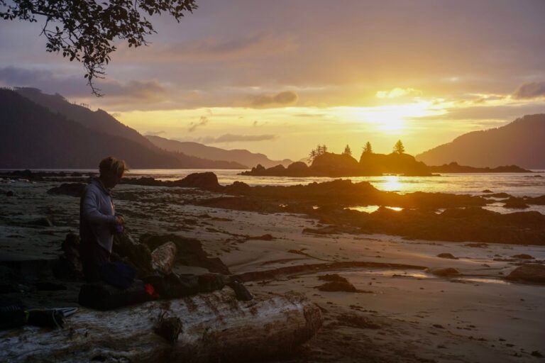 Person sitting on a rocky shore at sunset, with golden light reflecting on the water and mountains in the distance