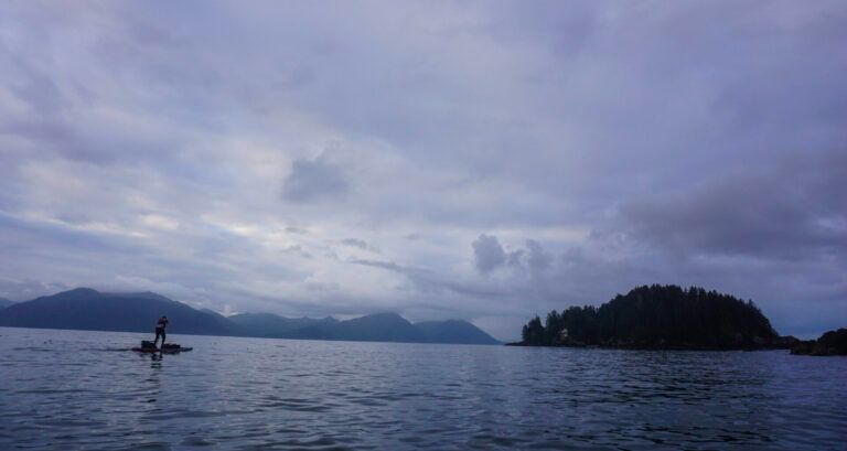Calm ocean at dusk with a lone paddleboarder, distant mountains, and a cloudy sky
