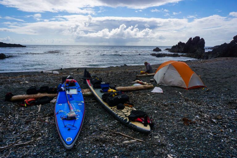 Kayaks and camping gear on a rocky beach beside an orange tent, with ocean and cloudy sky beyond.