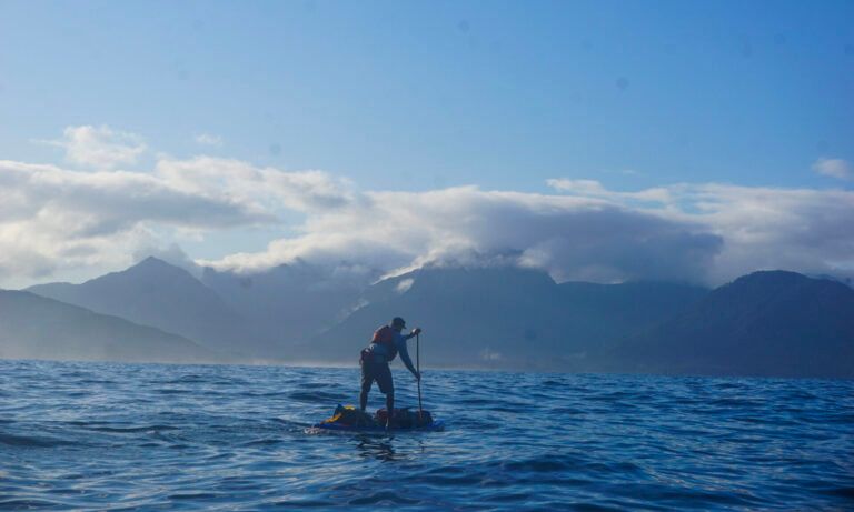 Person paddleboarding on choppy blue water with mountains and clouds in the background