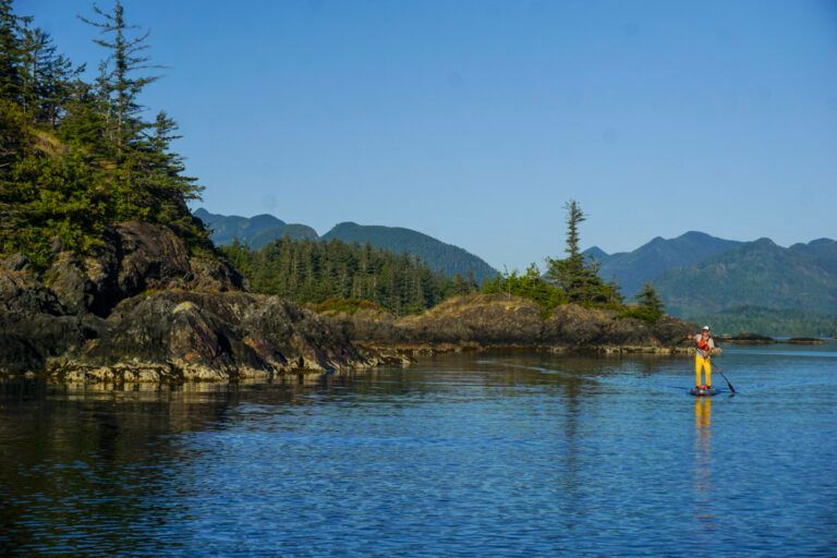 Calm blue lake with a small yellow kayak near rocky, forested shore and distant mountains under clear sky
