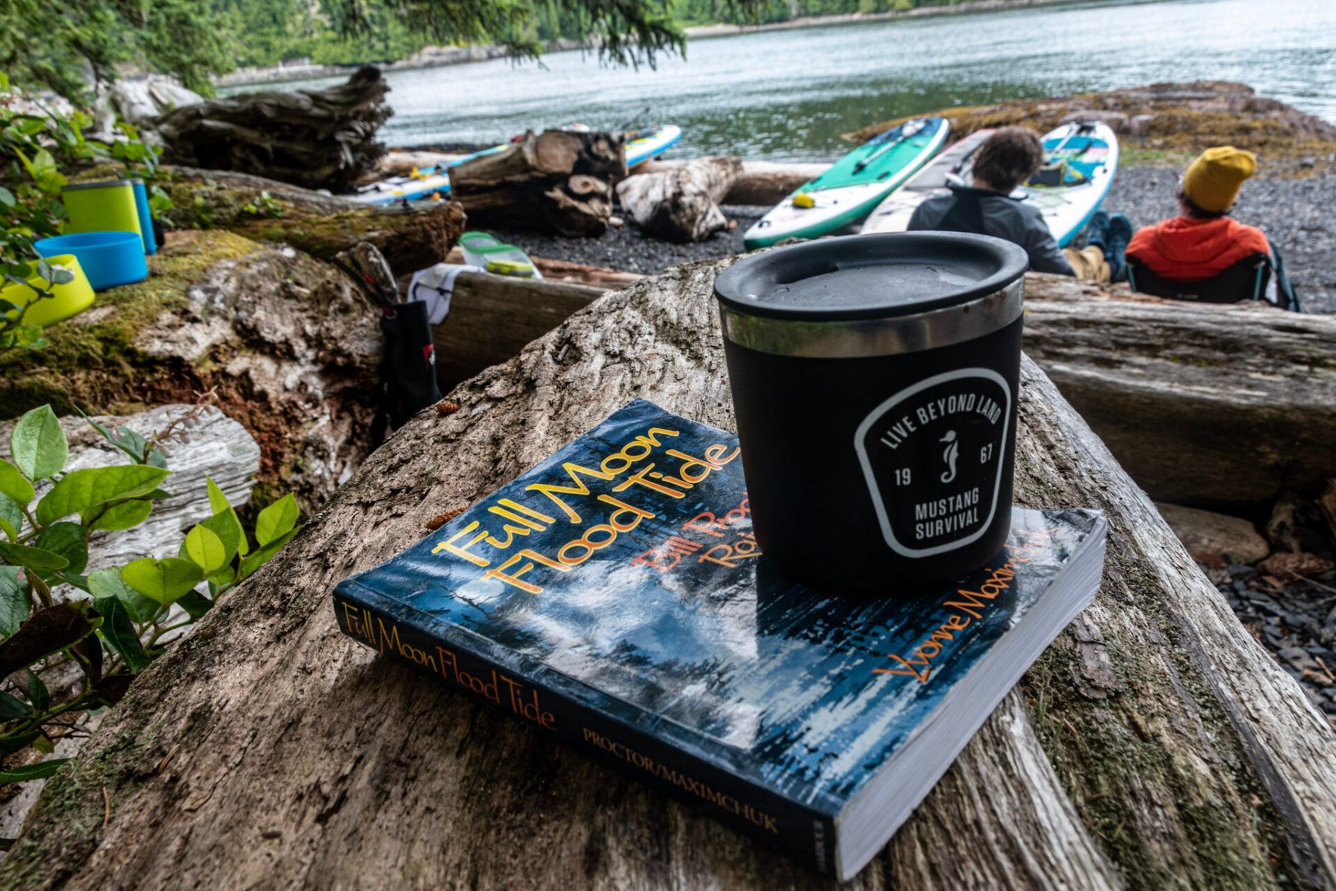 Coffee cup on a book on a rock by a river, with kayaks and a person in the background