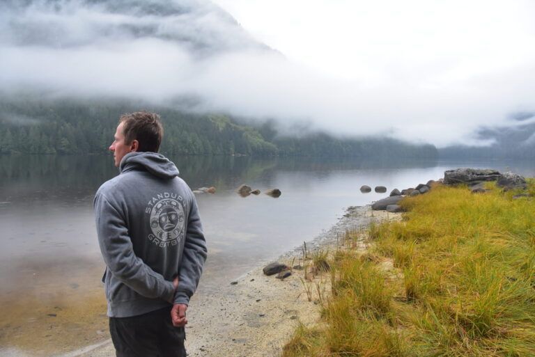 Person in gray hoodie standing by a misty lakeshore with forested hills and rocks.