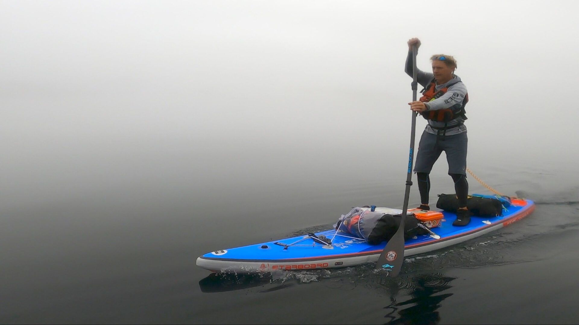 Person standing on a blue paddleboard in calm foggy water, paddling with a pole