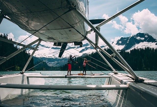 Two people stand on a boat deck under a large canopy, with mountains and water in the background.
