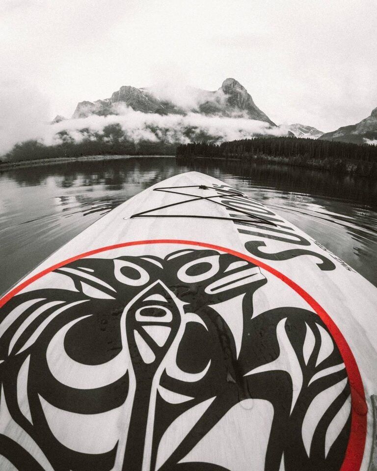 Kayak bow with black-and-white tribal design on a calm lake, mountains and clouds ahead