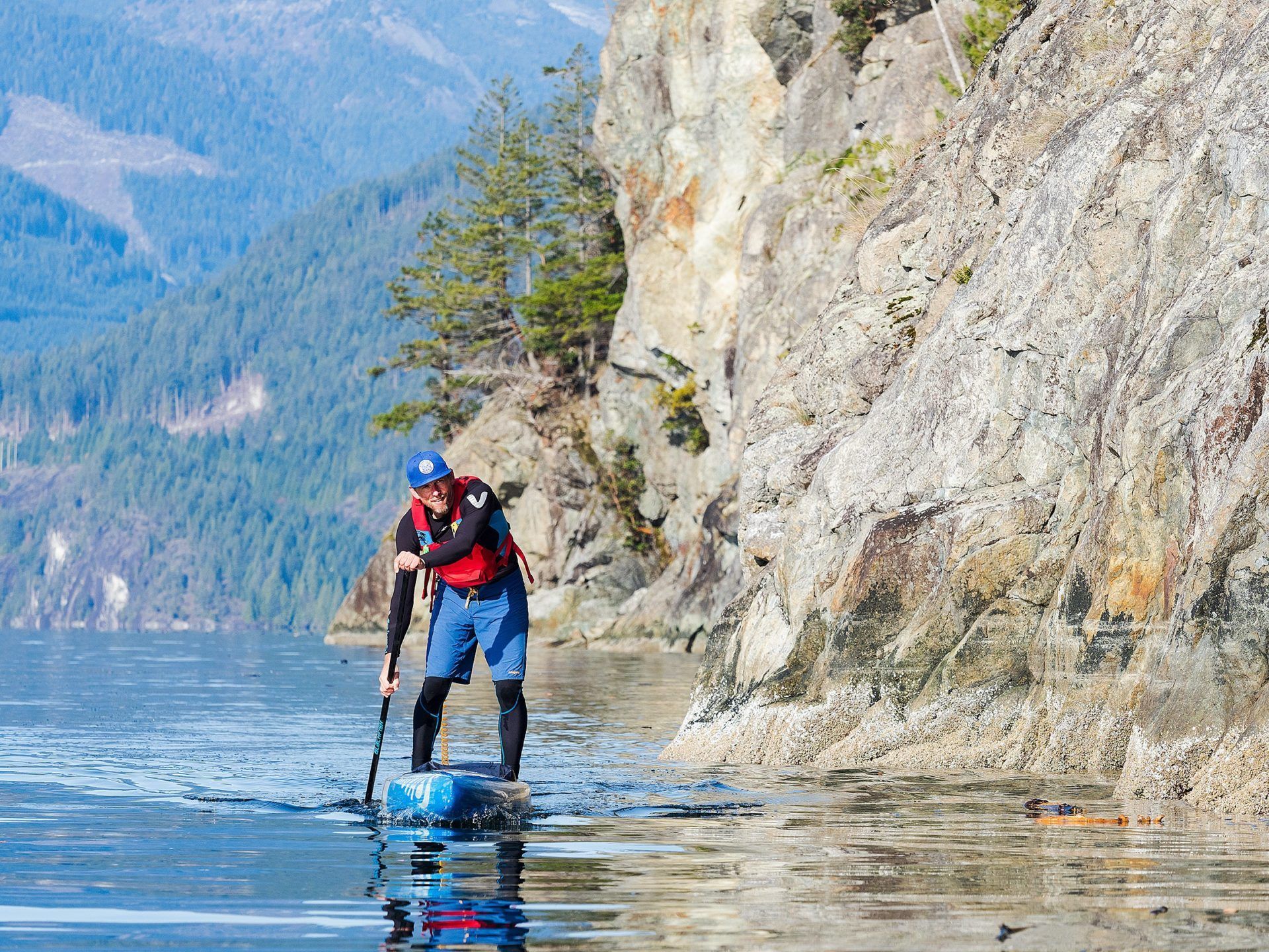 Person in red jacket paddling a stand-up board beside a rocky lakeshore with mountains in the background